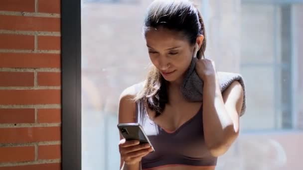 自宅のリビングルームに立って運動の休憩を取りながら 彼女のスマートフォンでメッセージを送信スポーティーな若い女性のビデオ — ストック動画