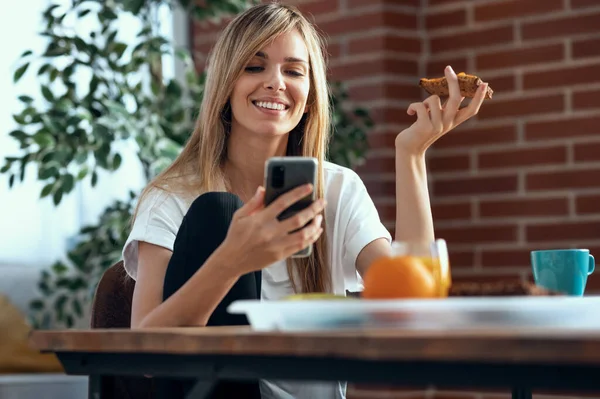 自宅のリビングルームで携帯電話を使いながら朝食を楽しむ笑顔の若い女性の肖像画 — ストック写真