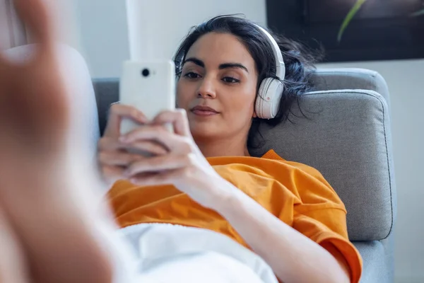 소파에 누워서 음악을 들으면서 핸드폰을 사용하는 자신감 여성의 — 스톡 사진