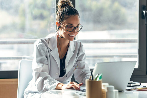 Shot of female doctor talking while explaining medical treatment to patient through a video call with laptop and earphones in the consultation.