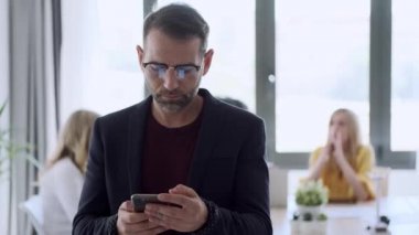 Yetişkin bir adamın kahve içerken cep telefonunu kullandığının videosu. Arka planda, iş arkadaşları ofiste çalışıyor..