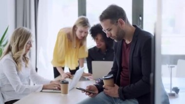 Yetişkin bir adamın kahve içerken cep telefonunu kullandığının videosu. Arka planda, iş arkadaşları ofiste çalışıyor..