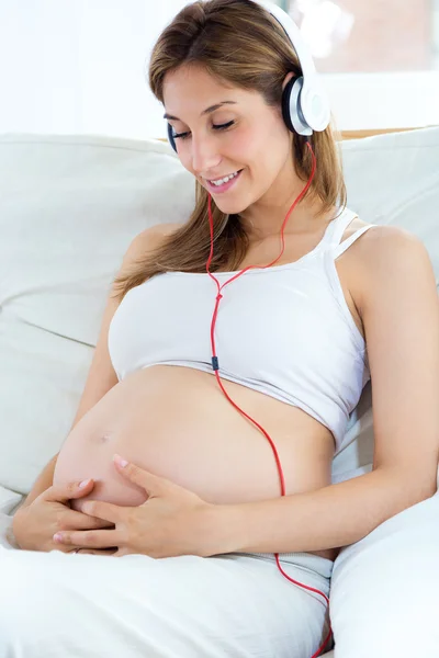 Zwangere vrouw zit op de bank en het luisteren naar muziek in koptelefoon. — Stockfoto