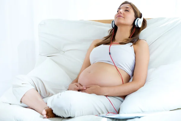 Gravid kvinna sitter på soffan och lyssnar musik i hörlurar. Stockfoto