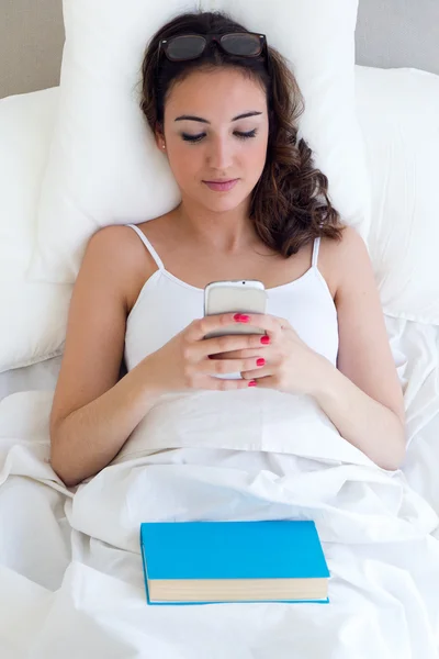 Hermosa mujer joven usando su teléfono móvil en la cama . — Foto de Stock