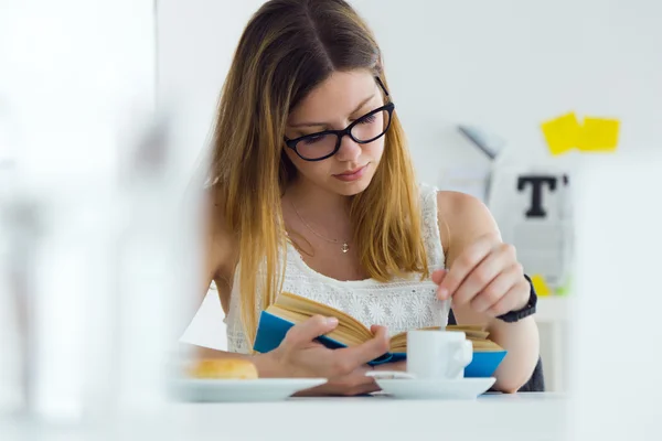 Ganska ung kvinna läser en bok och äter frukost hemma. — Stockfoto