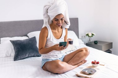 Yatakta otururken akıllı telefon kullanırken kahvaltıda kahve içen mutlu bir kadın.