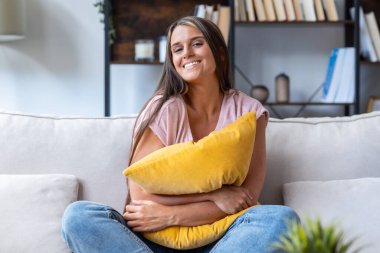 Evde koçun üzerinde oturup yastığa sarılmış gülümseyen bir kadının görüntüsü.