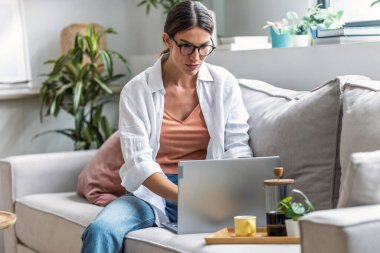 Evde kanepede otururken dizüstü bilgisayarla çalışan çekici bir kadının fotoğrafı.