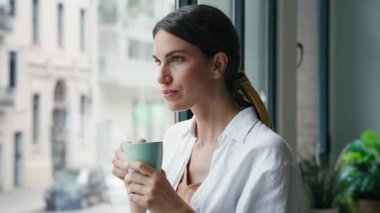 Güzel bir kadının bir fincan kahve içerken evinin penceresinin yanında dikilirken videosu..