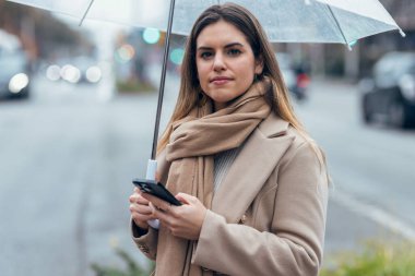 Güzel bir kadının, yağmurda şemsiyeyle yürürken kameraya bakarken telefonunu kullanması.