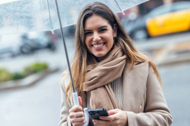 Güzel bir kadının, yağmurda şemsiyeyle yürürken kameraya bakarken telefonunu kullanması.