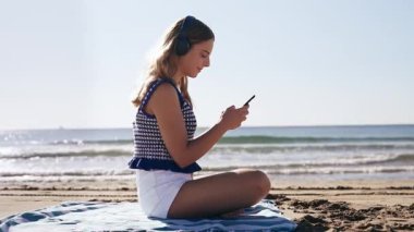 Deniz kenarında oturmuş kulaklıkla müzik dinlerken akıllı telefon kullanan tatlı bir kızın videosu.