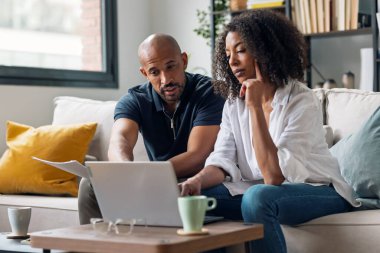 Yorgun çiftin evdeki kanepede otururken bazı fatura ve belgelere danışmak için dizüstü bilgisayarlarını kullanmaları.