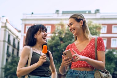 İki gülümseyen kadının şehirde gezerken akıllı telefon kullanması.