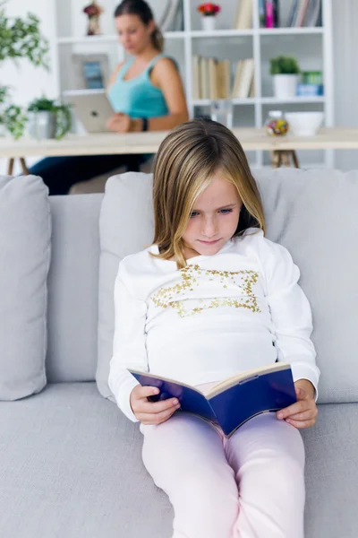 Criança lendo um livro enquanto sua mãe usa tablet digital em casa . — Fotografia de Stock