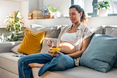 Hamile bir kadının evdeki kanepede otururken bir kase meyve salatası yemesi.