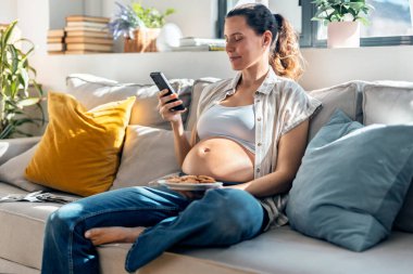 Hamile bir kadının evde kanepede oturmuş kurabiye yerken akıllı telefon kullanması.