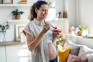 Evdeki oturma odasında bir kase çilek yiyen mutlu hamile kadın.
