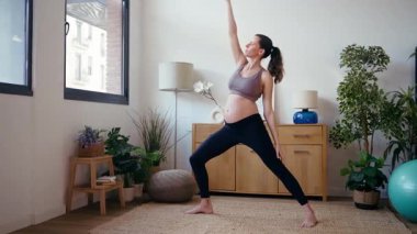 Eşofman giymiş hamile bir kadının evdeki oturma odasında yoga pozu verirken videosu.