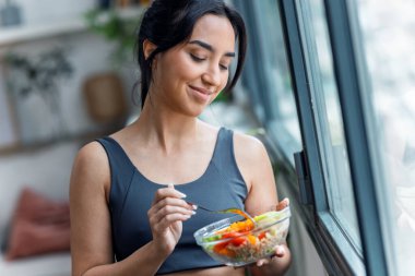 Sağlıklı bir kase yiyen sportif bir kadının evinde, mutfakta dikilirken çekilmiş fotoğrafı.
