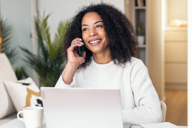 Evdeki mutfakta akıllı telefon kullanırken dizüstü bilgisayarla çalışan konsantre bir kadının fotoğrafı.