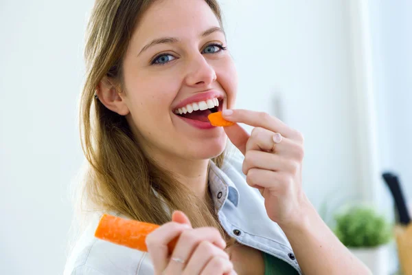 Mooie jonge vrouw eten wortel in de keuken. — Stockfoto