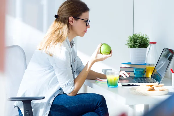 Mujer joven y bonita comiendo una manzana y trabajando en casa . — Foto de Stock