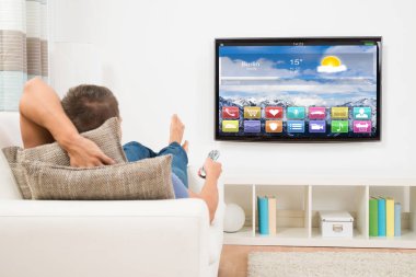 Televizyonun önündeki uzaktan kumandayı kullanarak koltukta uzanan genç adam.