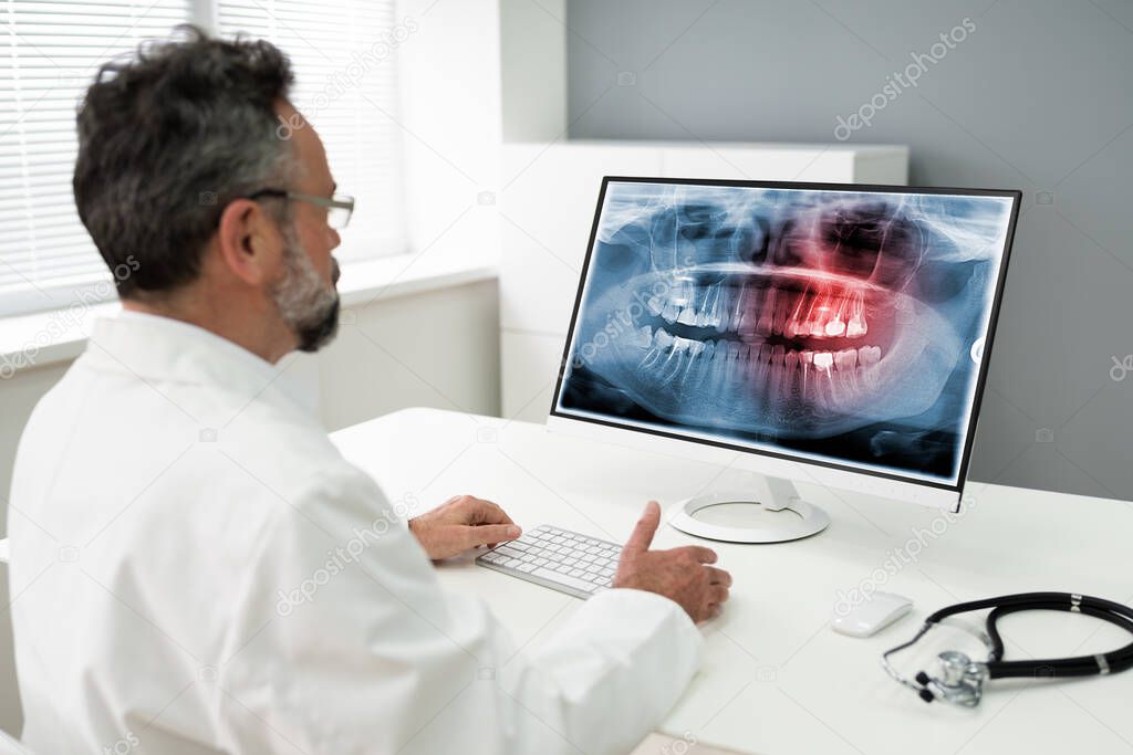 Mature Male Dentist Looking At Teeth X-ray On Computer In Clinic