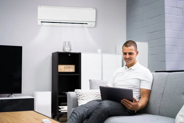 Jonge Gelukkige Man Zit Bank Met Behulp Van Airconditioning Thuis — Stockfoto