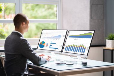 Businessman In Office At Desk Using Graphs On Computer Monitor