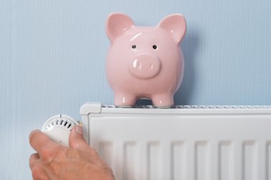 Man Holding Thermostat With Piggy Bank On Radiator clipart