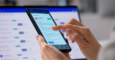 Young Businesswoman Managing Small Company Communications Using Smartphone And Laptop At Desk