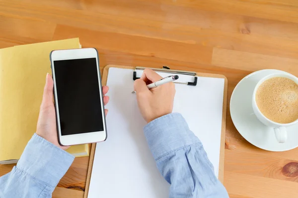 Woman writing on clipboard and using phone — Stock Photo, Image
