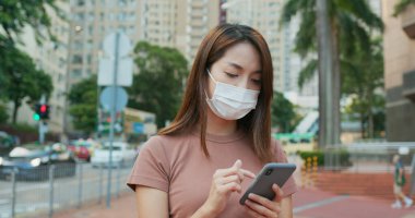 Kadın yüz maskesi takıyor ve açık havada akıllı telefon kullanıyor.