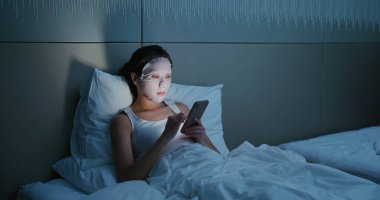 Kadın kağıt maske kullanıyor ve yatakta cep telefonu kullanıyor.