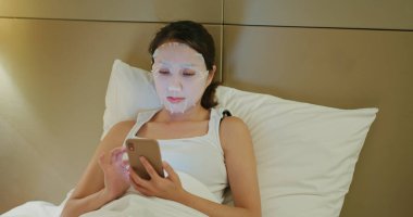 Kadın kağıt maske kullanıyor ve yatakta akıllı telefon kullanıyor.