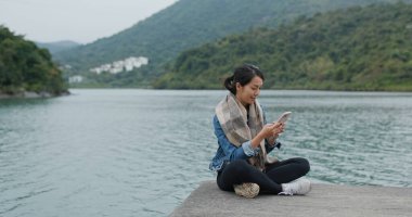 Kadın iskelede oturur ve akıllı telefon kullanır.