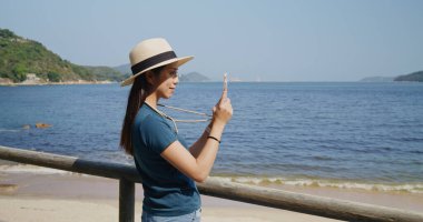 Kadın sahilde fotoğraf çekmek için cep telefonu kullanıyor.