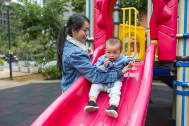 Genç anne bebeğinin parkta güvenle kaymasına yardım ediyor.