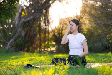 Genç bir kadın çimlerin üzerinde oturmuş, dışarıda dikkatli bir nefes alma egzersizi yapıyor.