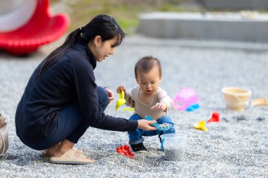 Anne, yeni yürümeye başlayan bebekle çakıl zeminde oynuyor.