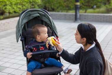 Anne dışarıda otururken bebeği muz ile besliyor.