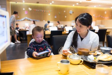 Anne bebeği restorana götürüyor, bebek de telefon izliyor.