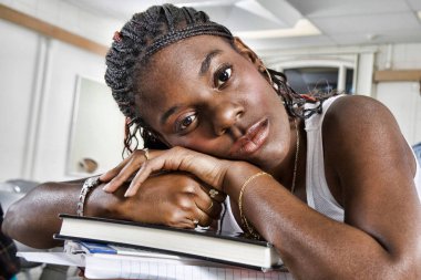 Closeup portrait of bored young African American student resting on stack of books in classroom clipart