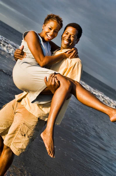 ビーチで女性を運ぶ陽気な若いアフリカ系アメリカ人の男性の写真 — ストック写真