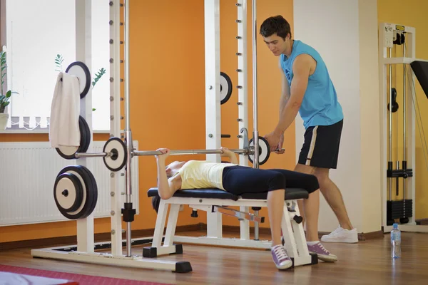 Young man helping a young woman lift a barbell on a bench — Stock Photo, Image