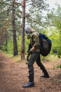 Gaz maskeli silahlı bir asker ormanda yürüyor ve düşmanı takip ediyor.