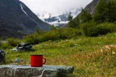 Çay ya da kahveli kırmızı bir kupa kar kaplı Altai dağlarının arka planında bir taşın üzerinde duruyor.